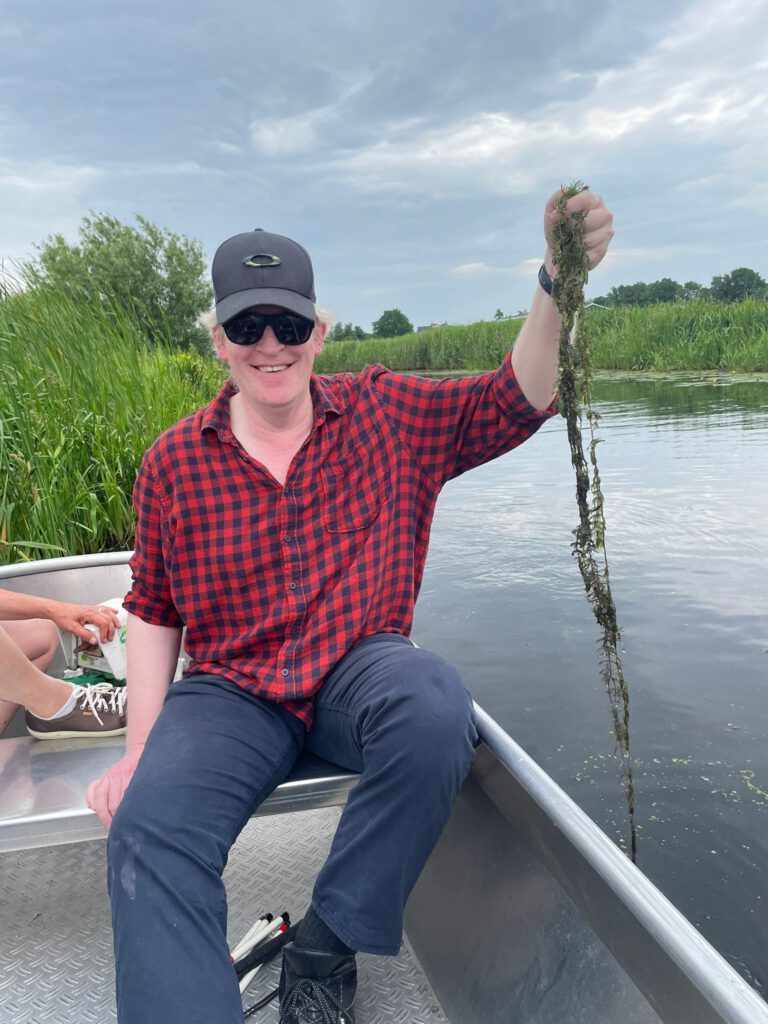 Richard houdt een sliert waterplanten omhoog terwijl hij in een bootje zit
